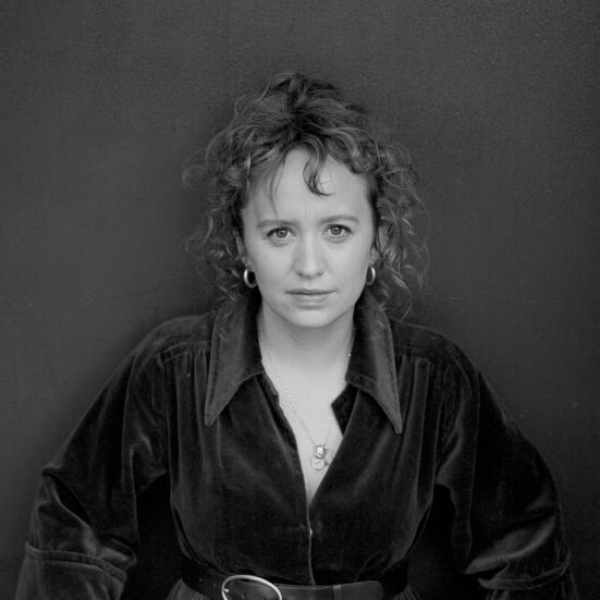 Sort-hvidt portræt af forfatteren Olga Ravn i mørk kjole med bælte, stående foran en mørk baggrund.